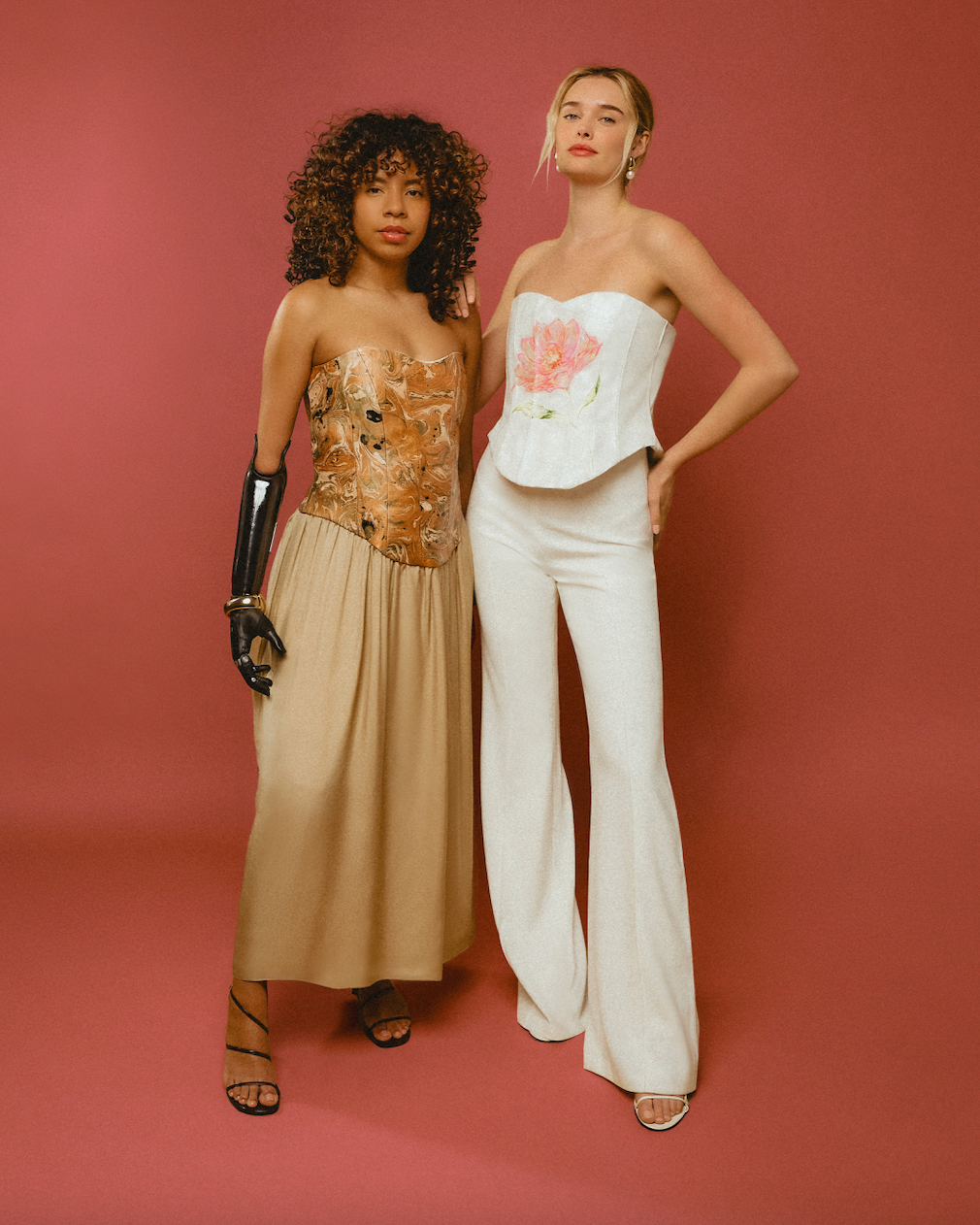 Two women posing against a pink background wearing stylish outfits.