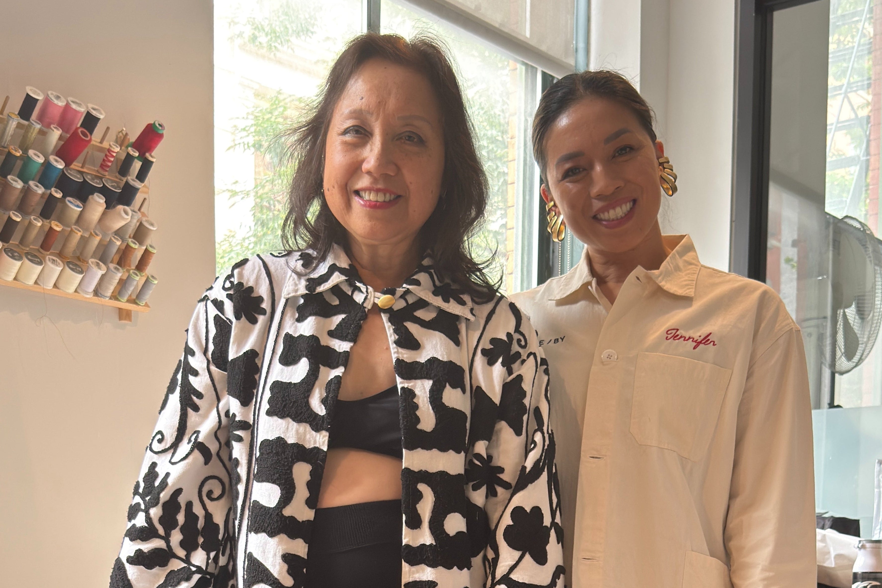 Two women standing indoors, one wearing a black and white patterned jacket, in a setting with shelves and windows.