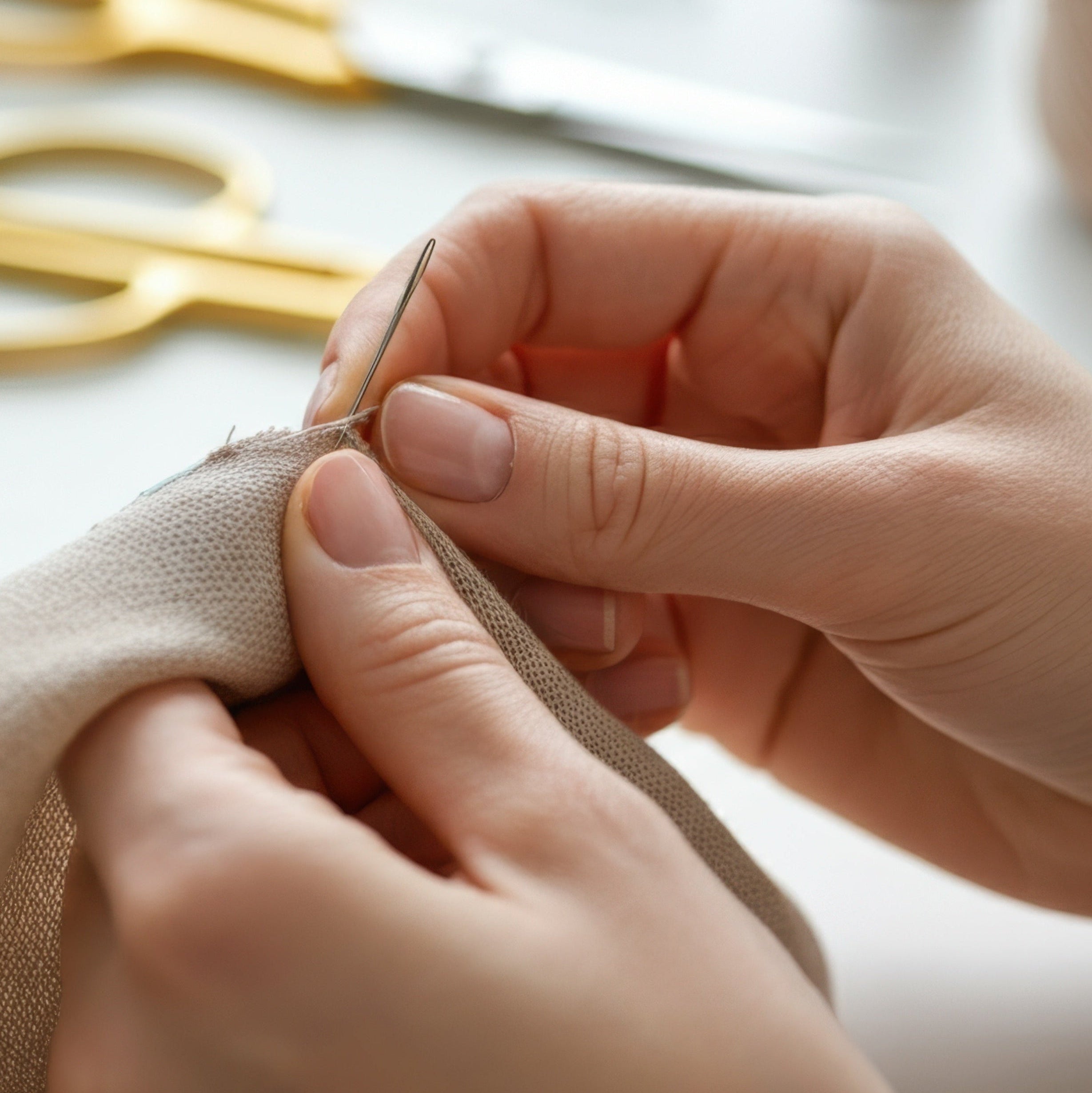 Close-up of hands sewing fabric with a needle and thread, scissors, and spools of thread in the background.