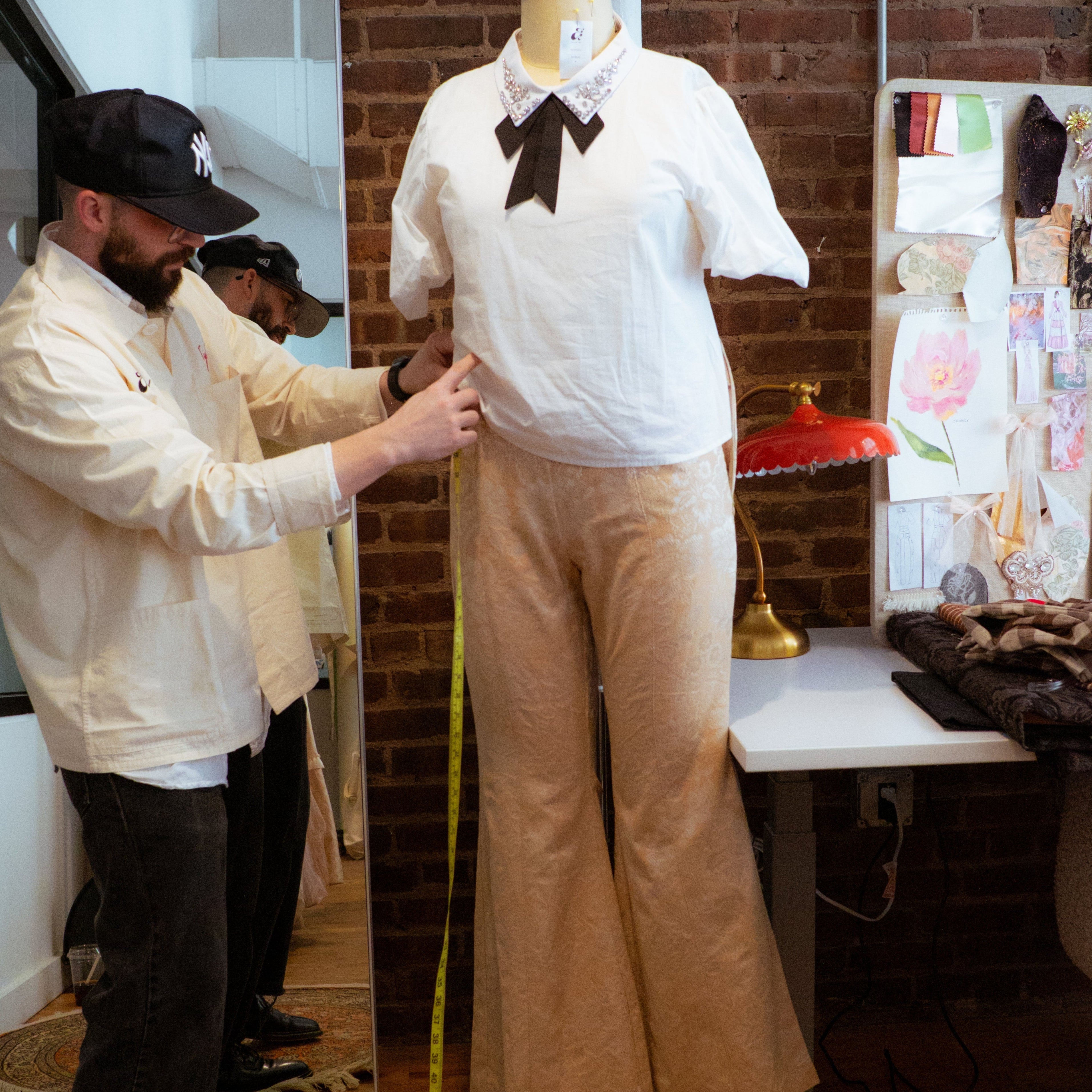 Person measuring a mannequin in a studio setting with brick walls and various items on shelves.
